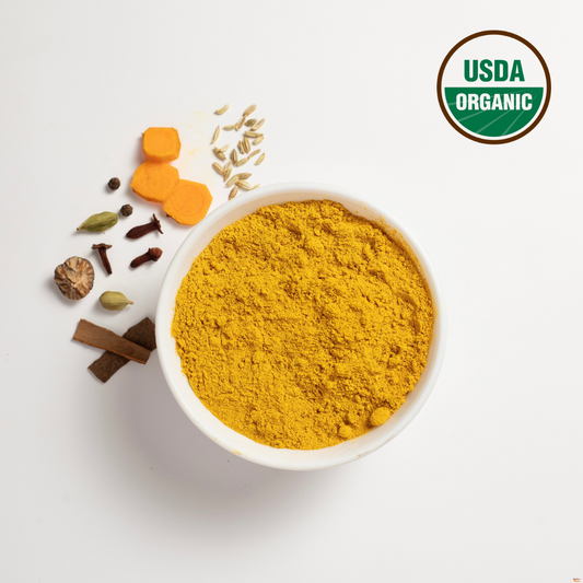 Organic turmeric blend is in a white bowl. Around the bowl are turmeric root, black pepper, fennel, cloves, cinnamon, cardamom and nutmeg. This turmeric spice blend is USDA organic as indicated by logo on the top right corner.