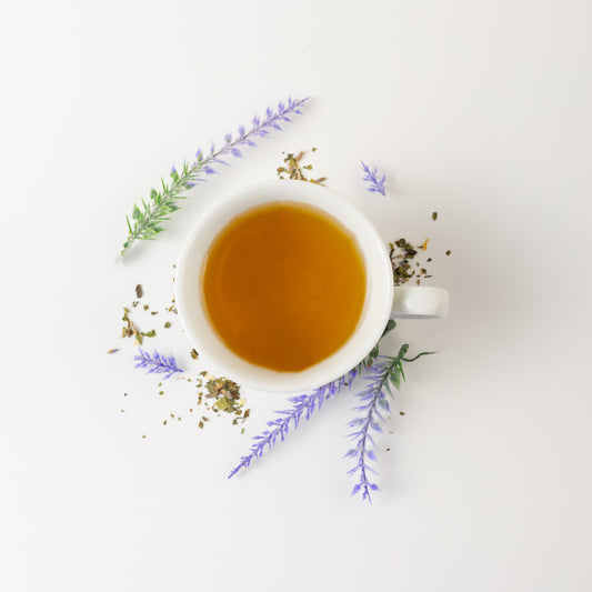 Lavender, the best herb for stress relief and lemon balm mint tea blend brewed in a white cup. Outside the cup is loose leaf lemon balm mint and lavender flowers