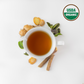 Organic Tulsi Ginger tea is brewed in a white cup. Outside the cup are holy basil leaves, organic ginger and organic licorice root. USDA organic certification for the tea is on the top right corner. 