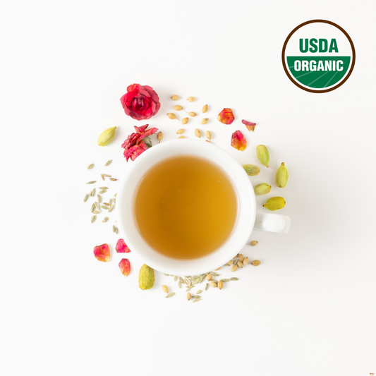 Organic Rose and Fennel tea blend is brewed in a white cup. Surrounding the cup are rose flowers, fennel, cardamom, coriander seeds which are pitta balancing herbs. On the right top corner is USDA organic logo indicating that this rose tea blend is certified organic.