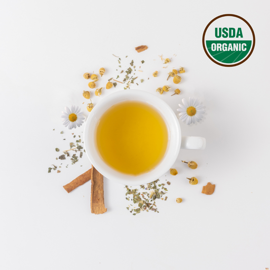 Brewed Organic chamomile and lemon balm tea blend in a white cup. Outside the cup are chamomile flowers, organic lemon balm mint and cinnamon. On the right top corner is USDA organic logo indicating that this night time chamomile tea blend is certified organic.