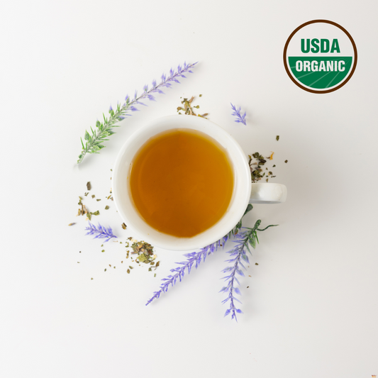 A white cup of lavender and lemon balm tea, which is the best tea for sleep. Outside the cup are lavender, the best her for anxiety and soothing lemon balm leaf. USDA organic logo is on the right top corner