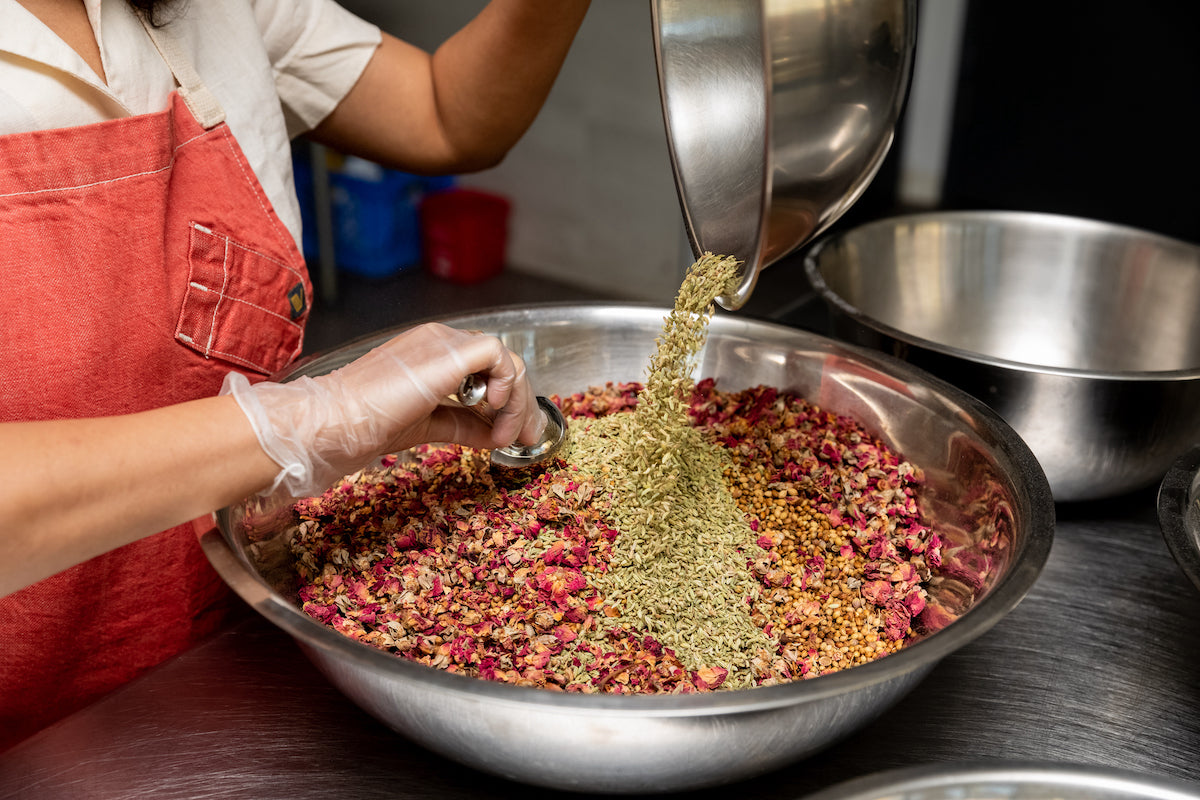 founder of ayursome foods, is making the organic rose tea blend with fennel and other anti inflammatory herbs. she is wearing a white shirt, a red apron, gloves and is blending the ayurvedic tea in a steel bowl with a steel scoop at a commercial kitchen.