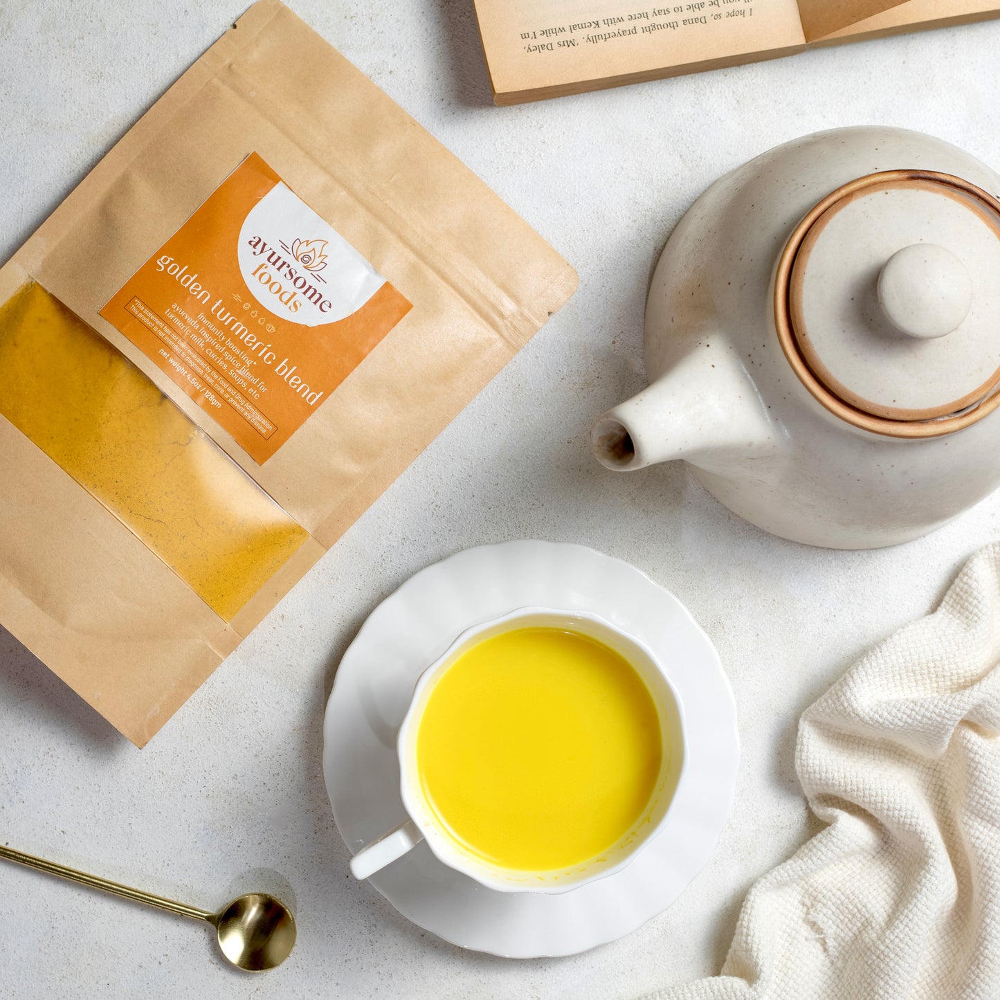 On a white background, there is pack of ayurveda turmeric spice blend, a cup of turmeric latte in a white cup, a golden color spoon, a white kettle, a white dinner napkin and a book. The golden turmeric spice blend is packed in a brown Kraft bag and is labelled in orange by Ayursome Foods.